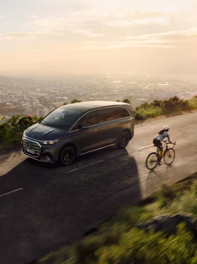 Mercedes Benz VLE 2026 - Sonnenaufgang, VLE fährt auf Straße, Stadt im Hintergrund zu sehen und im Vordergrund noch ein Fahrradfahrer