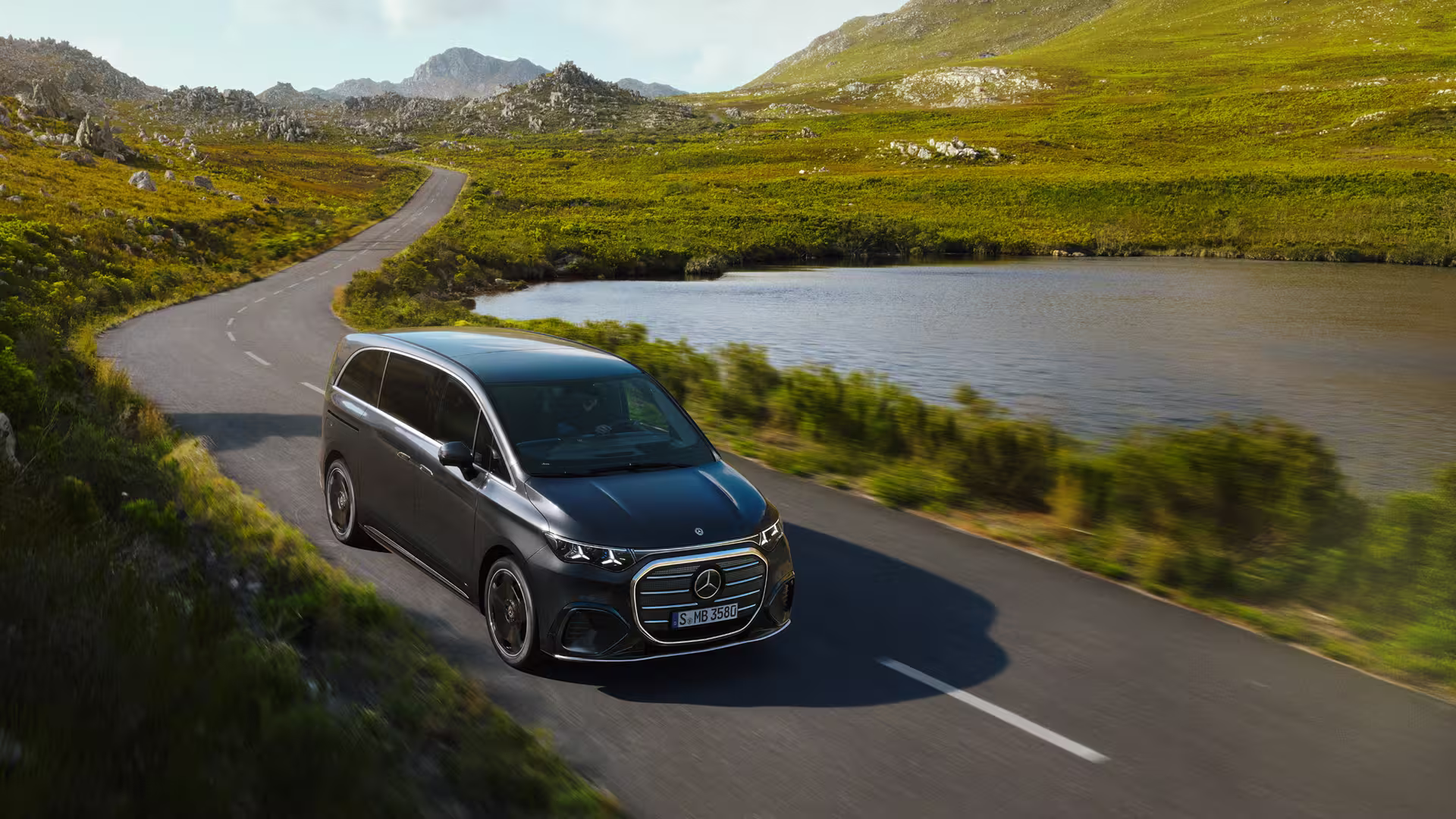 Mercedes Benz VLE 2026 - VLE fährt auf kurviger Straße, grüne Hügel im Hintergrund, sonnig